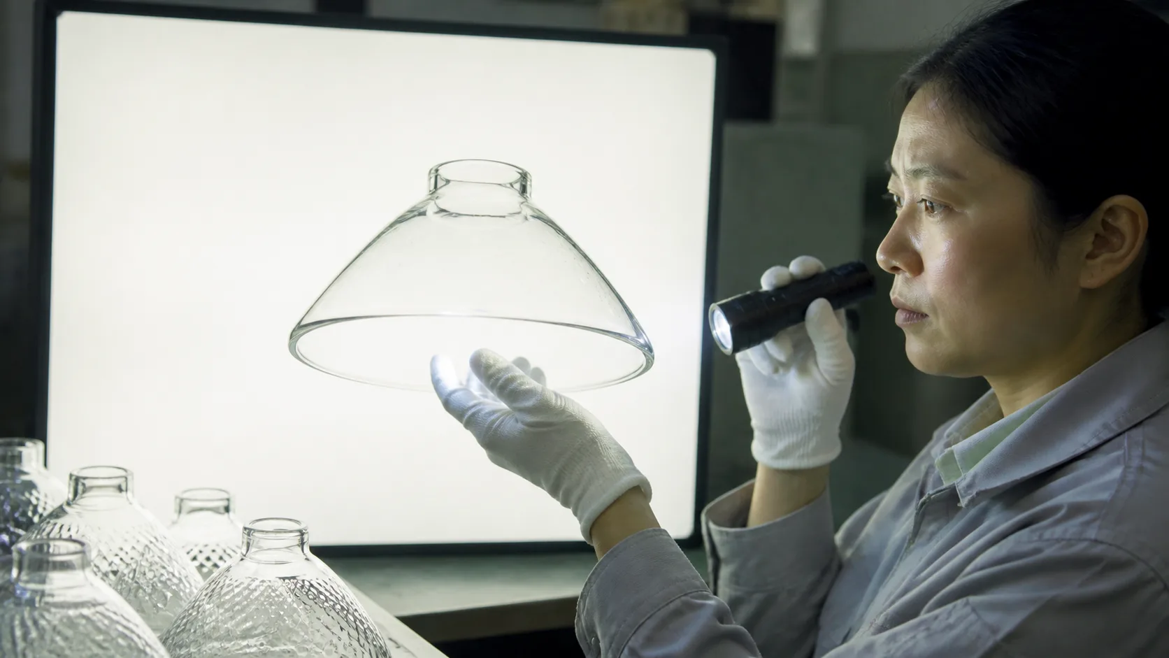 Coupelle de lampe en verre claire tenue devant un panneau d’inspection lumineux tandis qu’un inspecteur ganté vérifie les défauts de surface