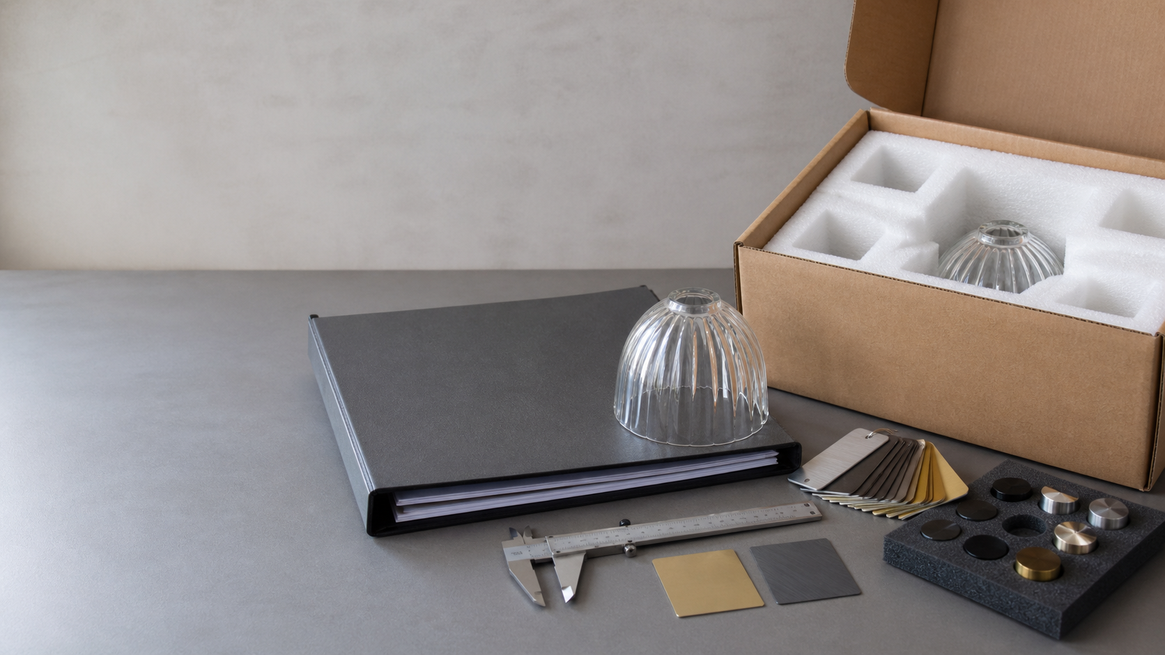 Ensemble complet prêt pour devis avec dossier de dessins, échantillon d’abat-jour en verre, pieds à coulisse, nuanciers de finitions et carton protégé sur un bureau d’approvisionnement